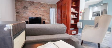Un salon avec un mur en briques, un téléviseur à écran plat, un canapé, une bibliothèque en bois et un fauteuil beige.