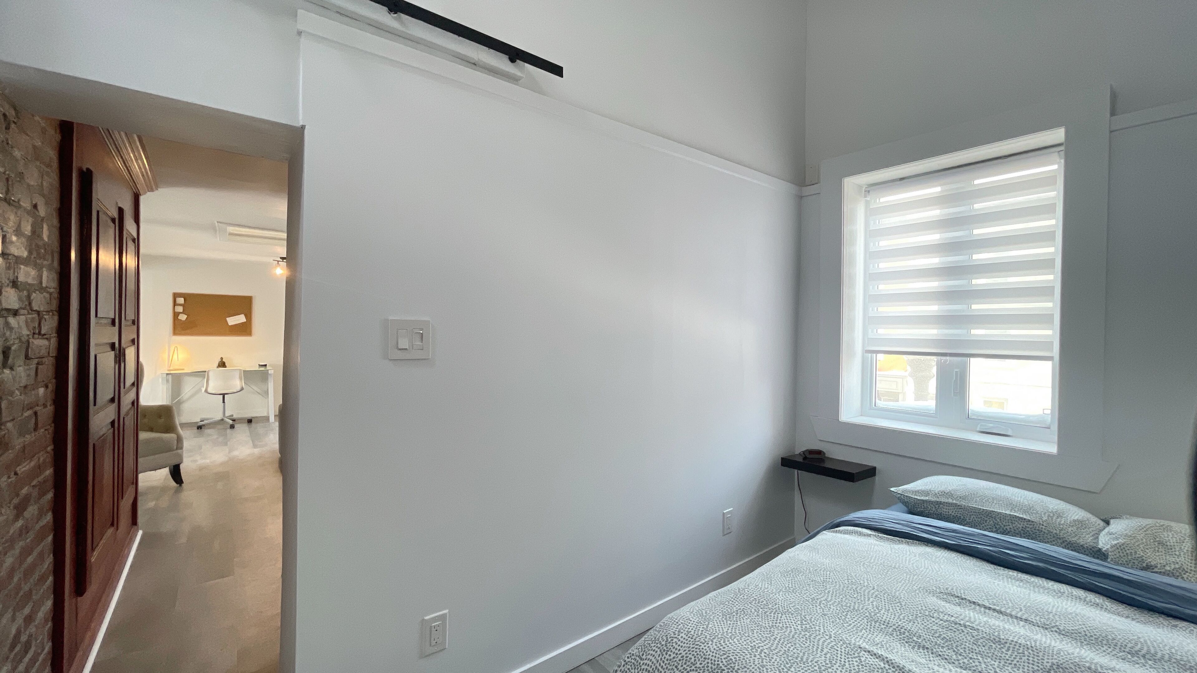 Un dormitorio con cama, una ventana con persianas, una mesita de noche y un enchufe en la pared.