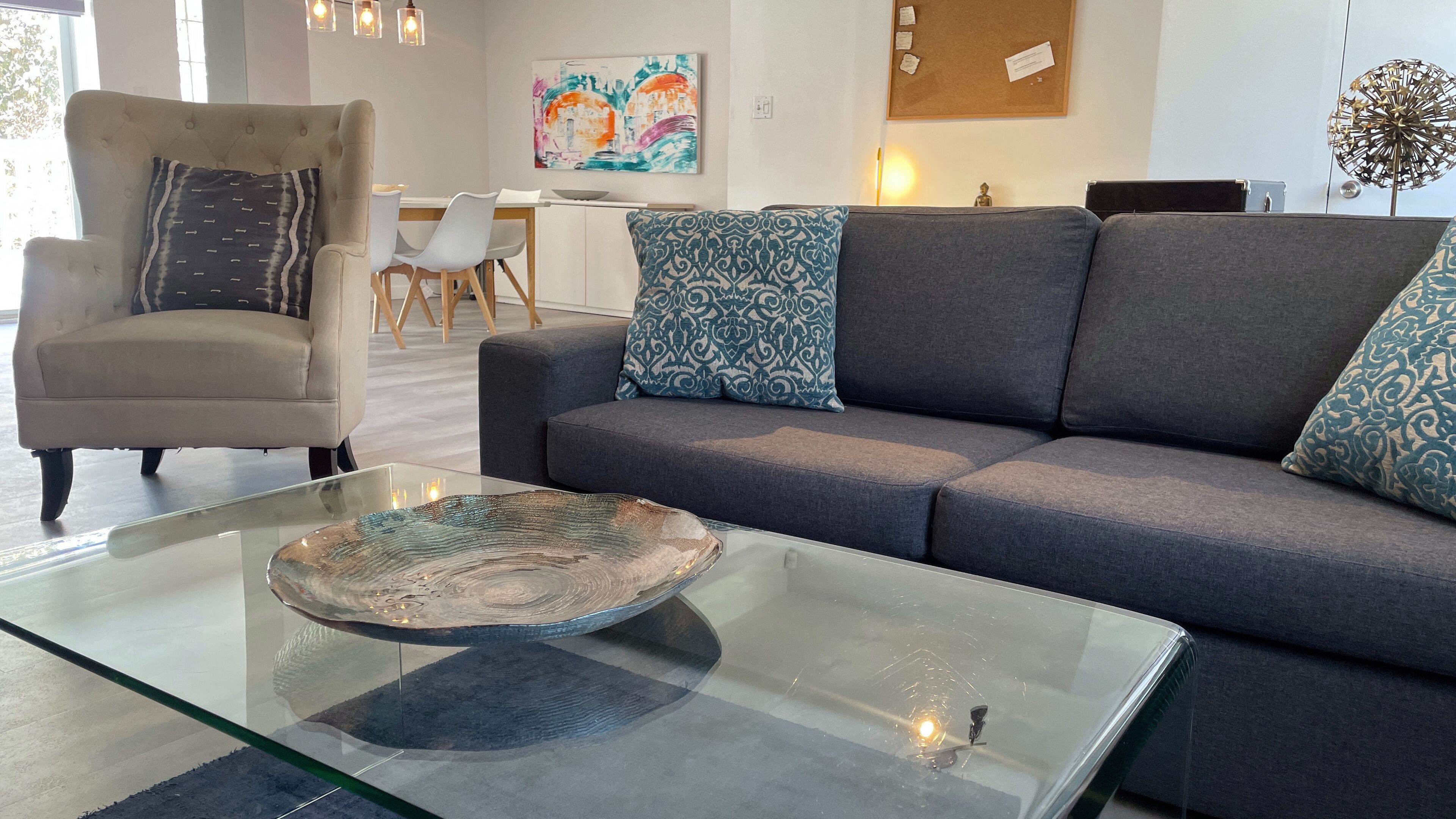 Una sala de estar moderna con un sofá gris, cojines estampados, una mesa de centro de vidrio con un cuenco decorativo, un sillón color beige y un área de comedor al fondo.