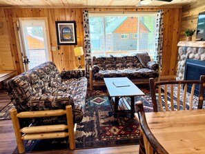 A cozy cabin interior with a wooden interior, a sofa, a table, and a view of a wooden house outside.