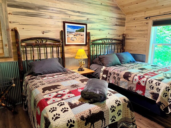 Ein Zimmer in einer Holzhütte mit zwei Betten, einem gerahmten Bild an der Wand und einem Fenster mit Blick auf Grün.