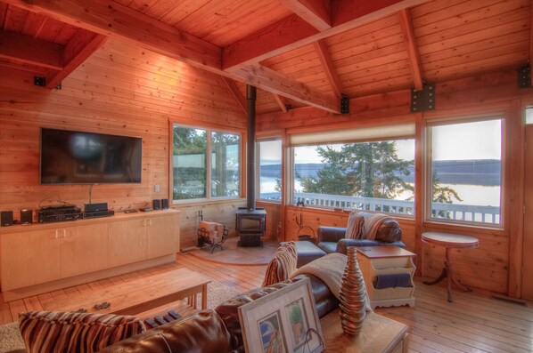 Un intérieur de chalet chaleureux, avec un décor en bois, une cheminée, un téléviseur et une vue sur l’extérieur.