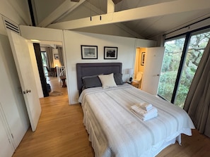 A bedroom with a large bed, two framed pictures on the wall, a bedside table with a lamp, and a view of greenery through a window.