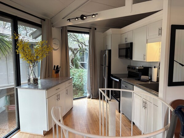 A modern kitchen with white cabinets, a stainless steel refrigerator, a microwave, and a dishwasher. There is a vase with yellow flowers on the island, and a clock on the wall. - Renovated Cottage, Walk to Beach, VIEWS, Golf Course, 2 BR/2 BA, 4 Bikes, Grill (Seabrook Island)