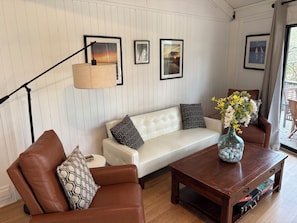 A living room with a white sofa, brown leather chairs, a wooden coffee table, and a floor lamp. There are framed pictures on the wall and a vase with flowers on the coffee table.