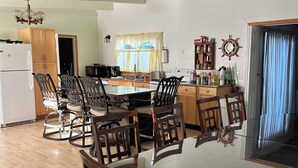 Un vaste espace de salle à manger avec une grande table et des chaises, une cuisine en arrière-plan et une vue sur l’extérieur par une fenêtre.
