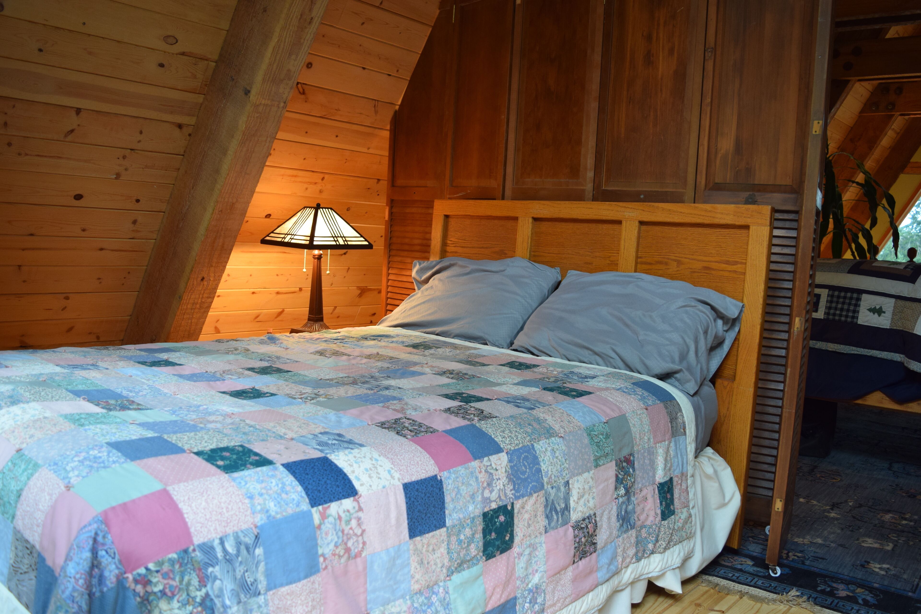 A bedroom with a wooden ceiling, a bed with a quilt, and a nightstand with a lamp.