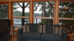 Un salon comprenant un canapé et un pouf, de grandes fenêtres donnant sur un lac et une forêt, ainsi que des meubles en bois.