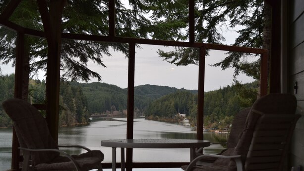 Une terrasse en bois avec deux chaises et une table, d’où l’on a vue sur un lac et des collines boisées.