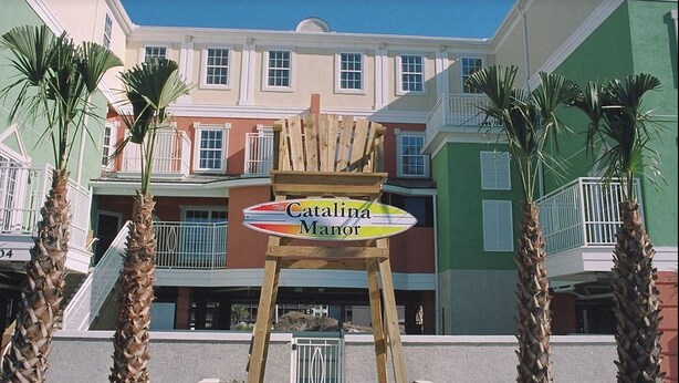 A wooden chair with a sign reading "Catalina Manor" in front of a building with balconies and palm trees. - In the HEART of North Myrtle Beach (North Myrtle Beach)
