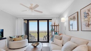Un salon avec un canapé, une table basse et un téléviseur. Il y a un balcon avec vue sur la plage.
