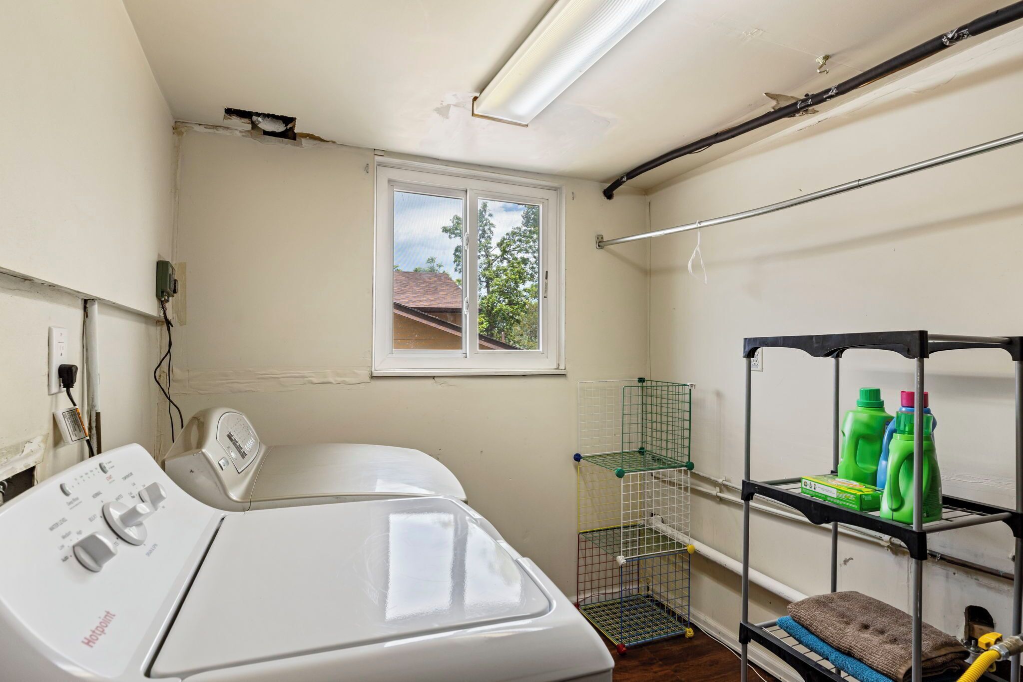 A laundry room with a washing machine, a drying rack, a window, and a shelf with cleaning supplies.