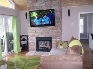 Cozy living room with a fireplace, flat-screen TV, and a view of lush greenery.