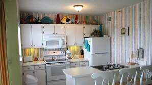 A kitchen with white cabinets, a microwave, a stove, a refrigerator, and a small island with chairs. - Inquiries are Beginning for Spring and Summer. Hear the Waves from  the Balcony. (Gulf Shores)
