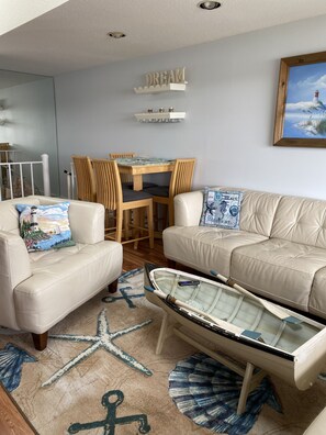 Ett vardagsrum med en beige soffa, ett soffbord med ett båtmotiv, en matta med ett sjöstjärnemönster och en tavla som föreställer en fyr.