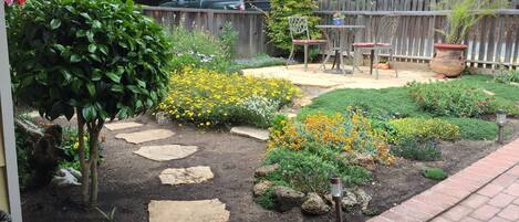 A garden with stepping stones, various flowering plants, a wooden fence, and a patio area with chairs and a table.