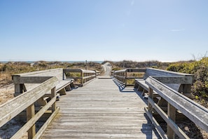 Ein hölzerner Steg führt zu einem Strand mit Dünen und spärlicher Vegetation.