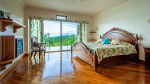 A bedroom with a wooden bed, a chair, a TV, and a balcony with a view of the countryside. - 2B / 2B IMPRESSIVE LAKE ARENAL, 3 POOLS, FULL KITCHEN (Tilaran)