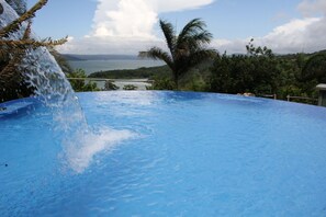 A swimming pool with a waterfall feature, surrounded by lush greenery and a view of the sea. - 2B / 2B IMPRESSIVE LAKE ARENAL, 3 POOLS, FULL KITCHEN (Tilaran)