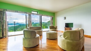 A spacious room with a large window offering a scenic view of a lush landscape. There are three beige armchairs and a wooden coffee table. A television is mounted on the wall. - 2B / 2B IMPRESSIVE LAKE ARENAL, 3 POOLS, FULL KITCHEN (Tilaran)