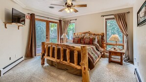 A bedroom with a rustic wooden bed, a ceiling fan, a TV mounted on the wall, and large windows with curtains.