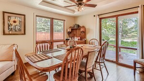 A dining area with a wooden table and chairs, a ceiling fan, and large windows with a view of greenery.
