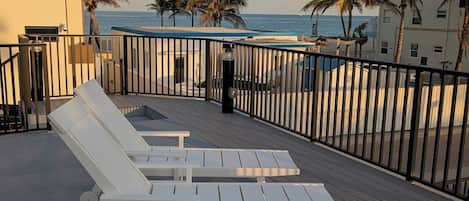 Una terraza en la azotea con sillones blancos, vista al océano, palmeras y un edificio con balcón.