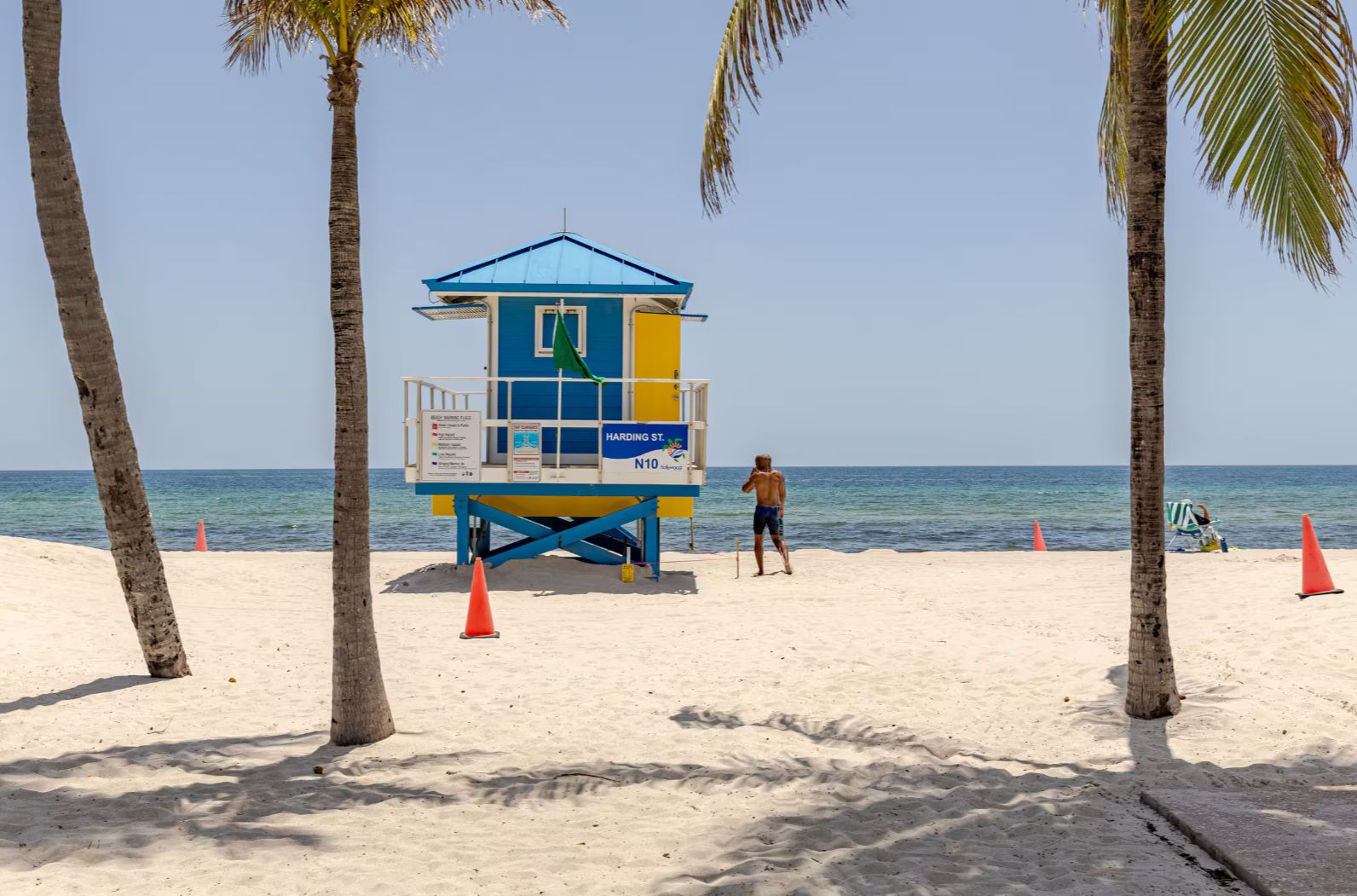 Une tour de surveillance, avec un toit bleu et des murs jaunes et bleus, située sur une plage de sable, avec des palmiers et l’océan en arrière-plan.