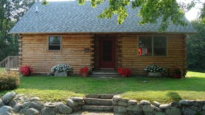 A log cabin with a red door, two windows, and a stone wall in front. - Cozy log cabin on Beech Hill. (Mount Desert)
