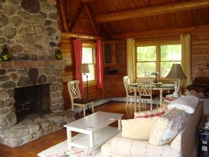 A cozy cabin interior with a stone fireplace, wooden walls, a dining area with a table and chairs, and a sofa with floral cushions. - Cozy log cabin on Beech Hill. (Mount Desert)