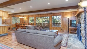 A spacious living area with a large gray sofa, wooden dining table, and chairs. There is a stone wall and a television on the right side.