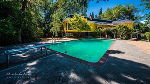 Una piscina con bordo in legno, circondata da alberi e un edificio giallo sullo sfondo.