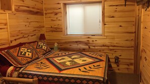 Een knus kamer in een chalet met houten inrichting, een bed met geborduurde dekens, een raam met zonwering en een klein nachtkastje met een zakdoosje.