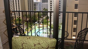 A balcony with a patterned sofa and chairs, overlooking a pool area with palm trees and buildings in the background. - Waikiki Banyan, Ocean-view 1BR apt: Full kitchen, FREE PARKING, NO RESORT FEES. (Honolulu)
