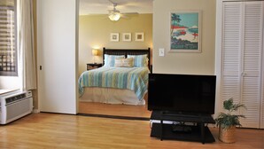 A hotel room with a bed, a television, a ceiling fan, a wall with a poster, and a door. - Waikiki Banyan, Ocean-view 1BR apt: Full kitchen, FREE PARKING, NO RESORT FEES. (Honolulu)