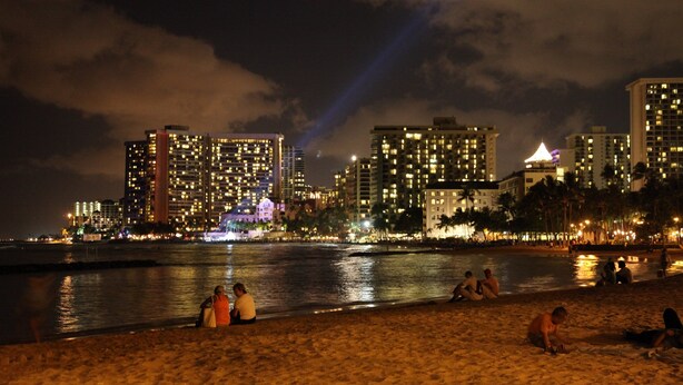 A beach with people sitting and walking along the shore, overlooking a city skyline with illuminated buildings. - Waikiki Banyan, Ocean-view 1BR apt: Full kitchen, FREE PARKING, NO RESORT FEES. (Honolulu)