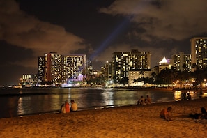 A beach with people sitting and walking along the shore, overlooking a city skyline with illuminated buildings.