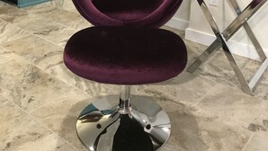 Een paarse fluwelen fauteuil met een metalen voet in een kamer met betegelde vloer, een keukenhoek en een aan de muur bevestigde televisie.