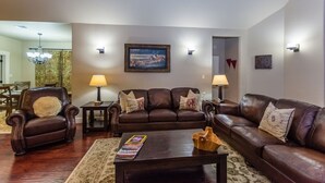 Un salon avec un canapé en cuir marron, deux fauteuils, une table basse et deux petites tables d’appoint avec des lampes. Un tableau orne le mur et un lustre se trouve au fond de la pièce.