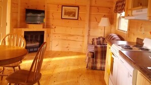 L’intérieur chaleureux d’un chalet comprend une table à manger, des chaises, une cheminée, un canapé, un coin cuisine avec une cuisinière et un ventilateur de plafond.