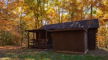 A rustic cabin with a porch and a covered seating area, surrounded by a forest with autumn foliage.