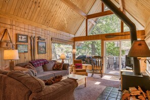 A cozy cabin interior with a wooden ceiling, a fireplace, and a large window overlooking a forest. - Icicle Riverhaus-Cozy Riverfront Cabin Leavenworth (Leavenworth)