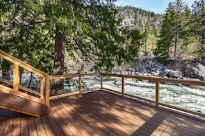 A wooden deck with a railing overlooking a river, surrounded by trees and a rocky hillside.
