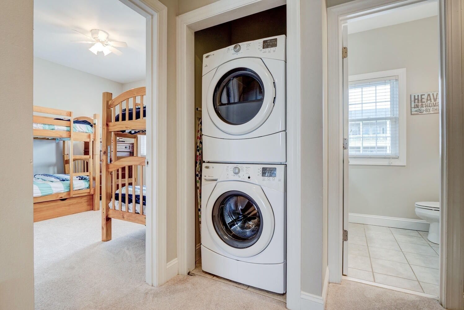 A laundry room with two stacked front-loading washing machines, a wooden bunk bed, and a small window with blinds.