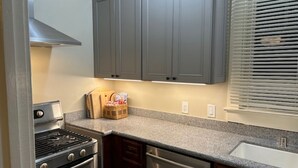 A kitchen with grey cabinets, a stainless steel stove, a dishwasher, and a window with blinds. - Lavender House Cottages, Classic Elegance in a Country Cottage (Point Reyes Station)