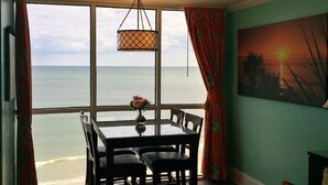 A dining area with a view of the sea, a table with chairs, a hanging light fixture, and a painting on the wall. - "Chasing Waves"-Oceanfront Luxury King Condo w/ Kitchen, Living Room & that View (Myrtle Beach)