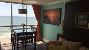 A room with a view of the ocean, a dining table with chairs, a sofa with a cushion, and a painting on the wall. - "Chasing Waves"-Oceanfront Luxury King Condo w/ Kitchen, Living Room & that View (Myrtle Beach)