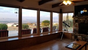 A room with large windows offering a view of a mountainous landscape. There is a stone fireplace and a television mounted above it. - Little Park Road Retreat - Hot Tub with a View (Grand Junction)