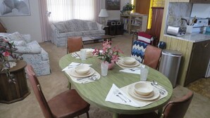 Un coin repas avec une table verte dressée pour quatre personnes, entourée de chaises marron. Un arrangement floral est posé sur la table, et une bibliothèque se trouve à l’arrière-plan.