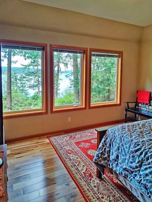A bedroom with a large bed, a red chair, and three windows offering a view of trees and a body of water.